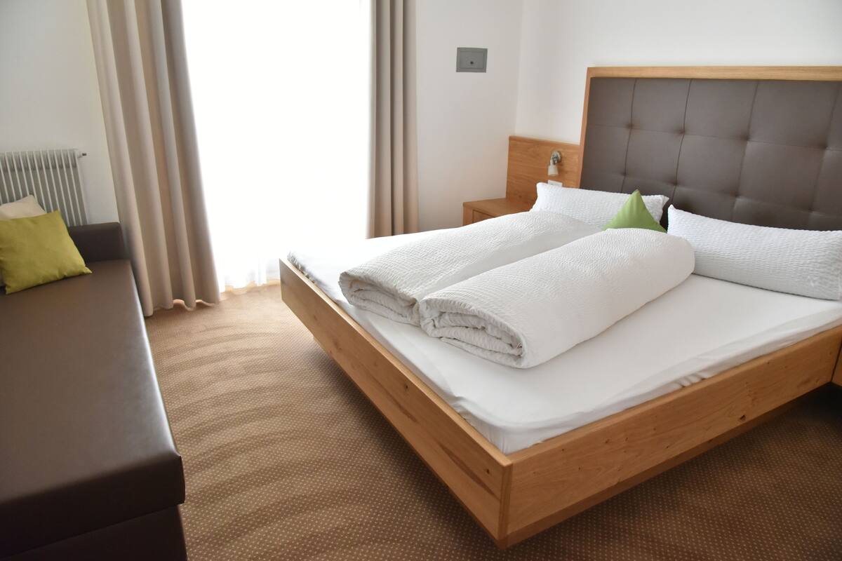 A neatly made bed with white linens and pillows, a wooden headboard, a wooden nightstand with a lamp, and a view of the outdoors through curtains.
