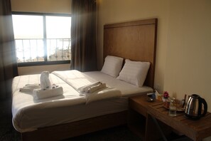 A hotel room with a large bed, two pillows, a wooden headboard, a nightstand with a kettle, cups, and a glass. - Holiday aqaba hotel (Aqaba)
