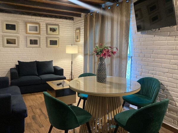 A cozy living room with a round glass table, green chairs, a black sofa, and a TV mounted on the wall.