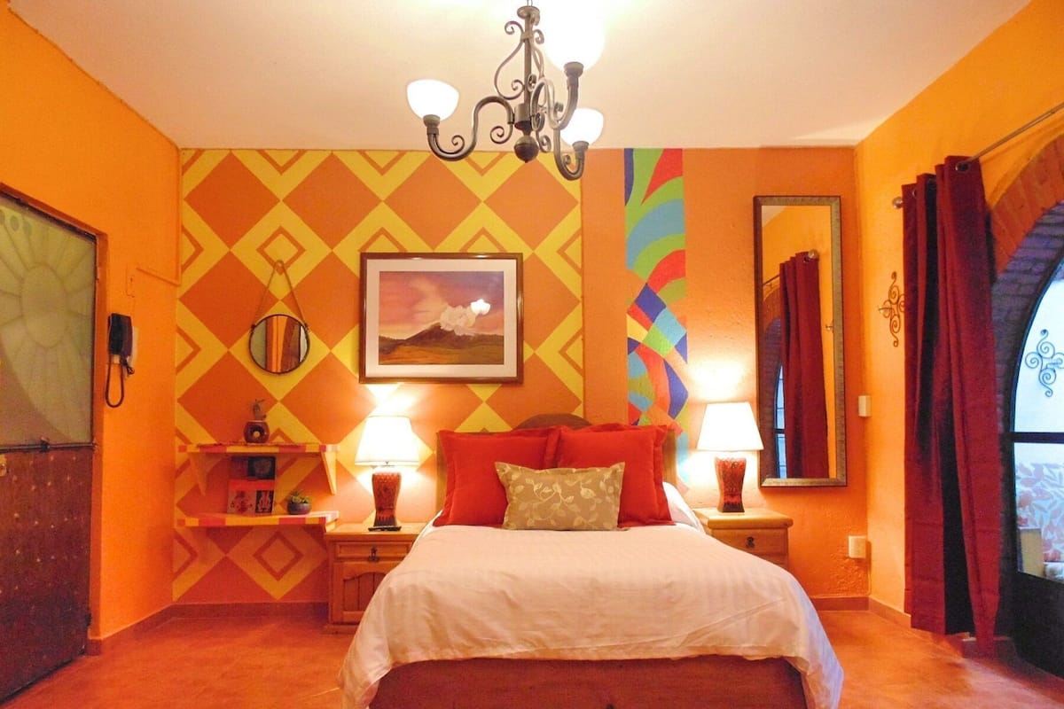 A bedroom with a bed, two bedside tables, a chandelier, a mirror, and a colorful wall pattern.