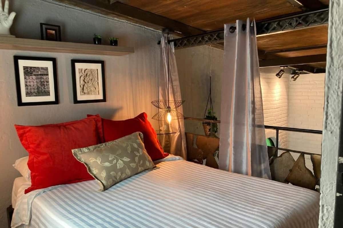 A bed with striped bedding, red and beige pillows, a wall-mounted light fixture, and framed artwork.