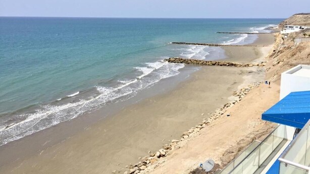 A beach with a rocky breakwater, clear blue water, and a distant view of a white building. - Golden Mar Hostal (Manta)