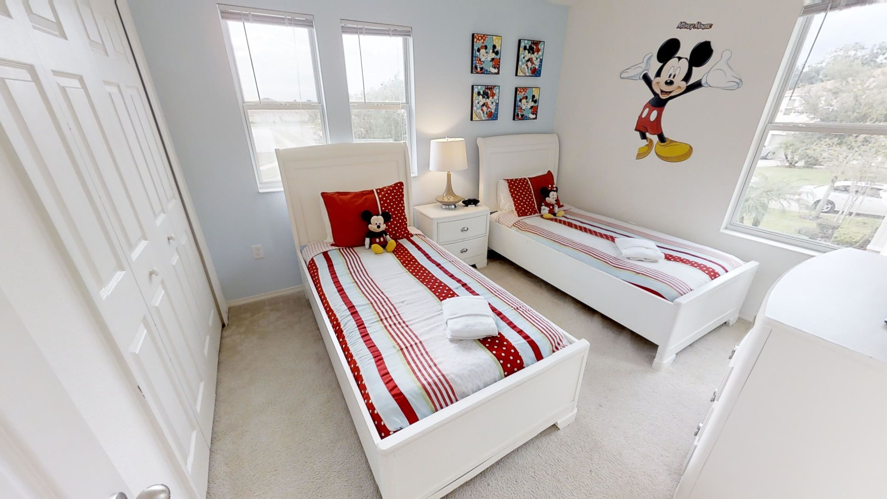 Une chambre avec deux lits simples, une table de chevet avec une lampe, un autocollant mural à l’effigie de Mickey Mouse et une fenêtre avec des stores.