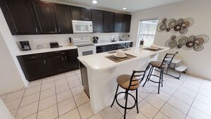 A modern kitchen with dark cabinets, a white countertop, a central island with stools, and decorative wall art. - Crystal Cove Resort (Kissimmee)