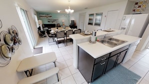 A modern kitchen with white cabinetry, a central island with a sink, and a dining area with a table and chairs. - Crystal Cove Resort (Kissimmee)
