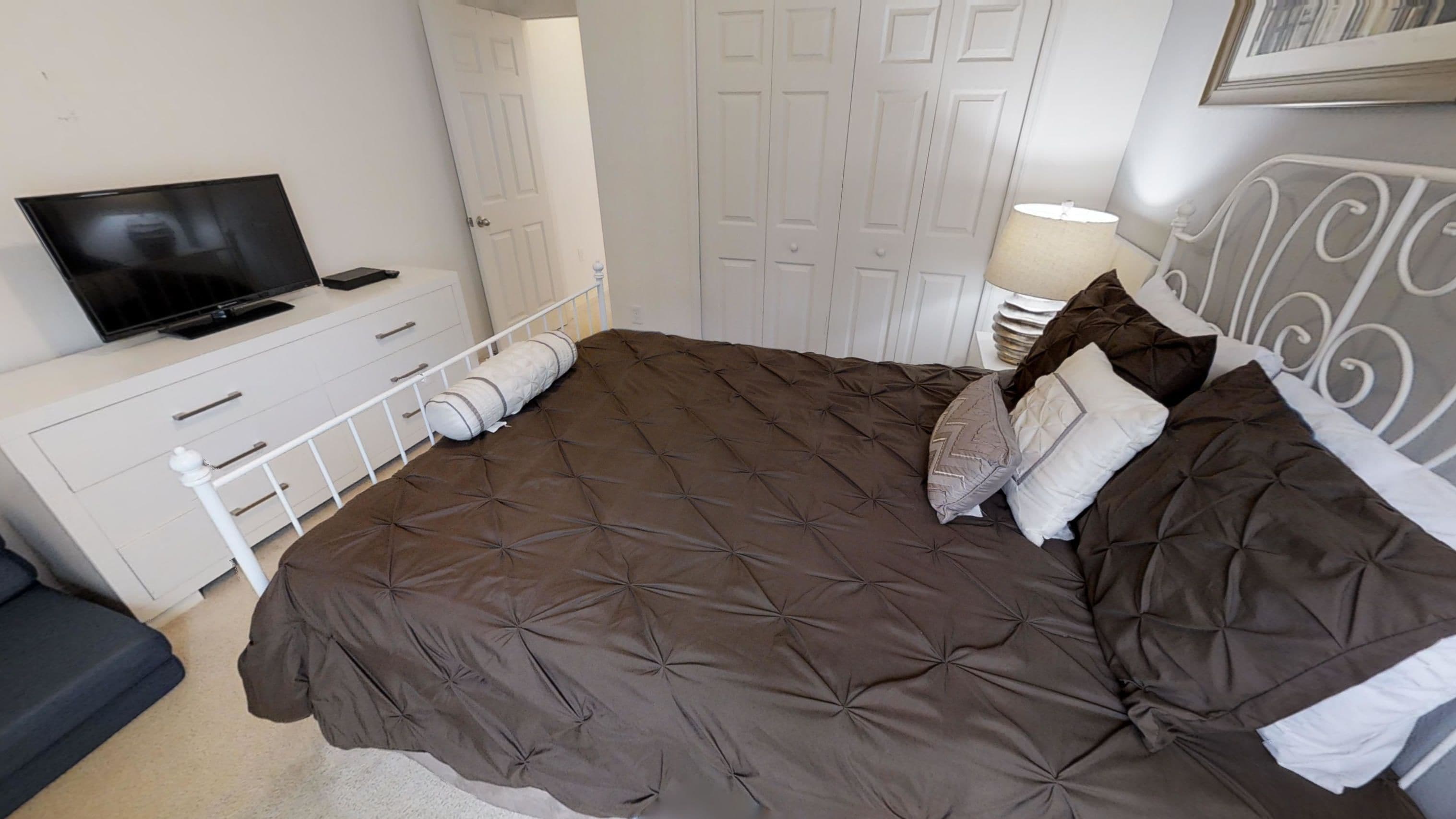 Ein ordentlich eingerichtetes Schlafzimmer mit einem großen Bett, einem Fernseher auf einer weißen Kommode und einer Tür im Hintergrund.