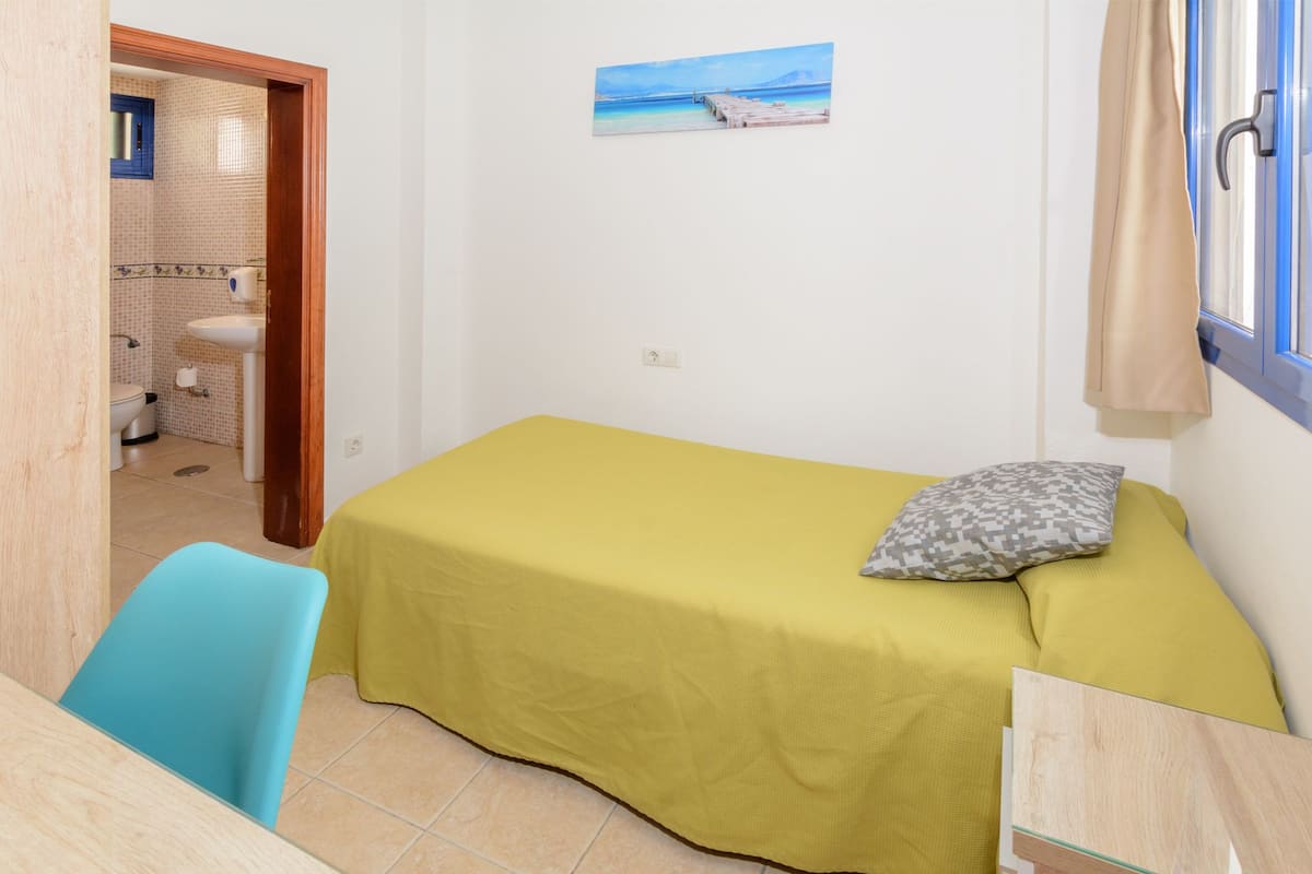 A small, single-bed room with a yellow bedspread, a pillow, a blue chair, a window with curtains, and a painting of a beach scene on the wall.