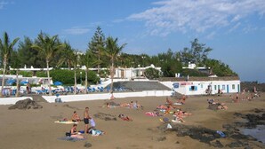 A beach with people sunbathing and swimming, palm trees, and a white building with a blue door. - Pensión MAGEC (Tías)