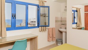 A hotel room with a desk, chair, bed, bathroom, and a view of the sea through blue-framed windows. - Pensión MAGEC (Tías)