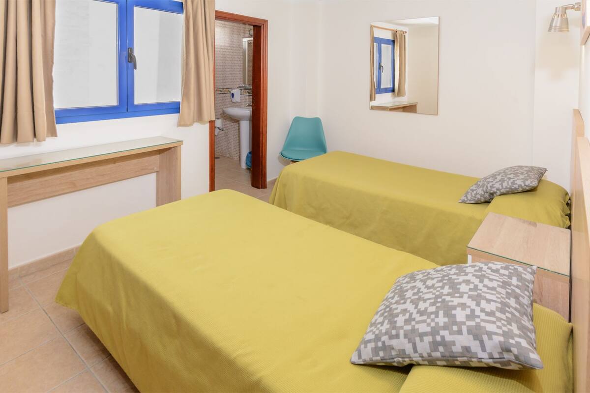 A room with two beds, a desk, a chair, a mirror, and a bathroom visible through an open door.