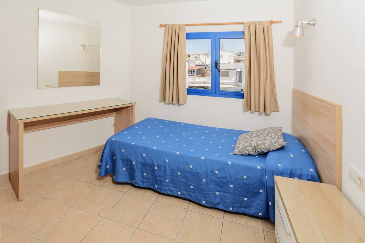 A bedroom with a bed, a desk, a window with curtains, and a view of buildings outside.