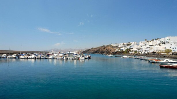 A marina with boats docked, a clear blue sky, and white buildings on a hillside. - Pensión MAGEC (Tías)
