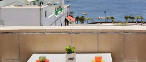 A balcony with a table set for two, overlooking a coastal view with the sea and a distant shoreline.