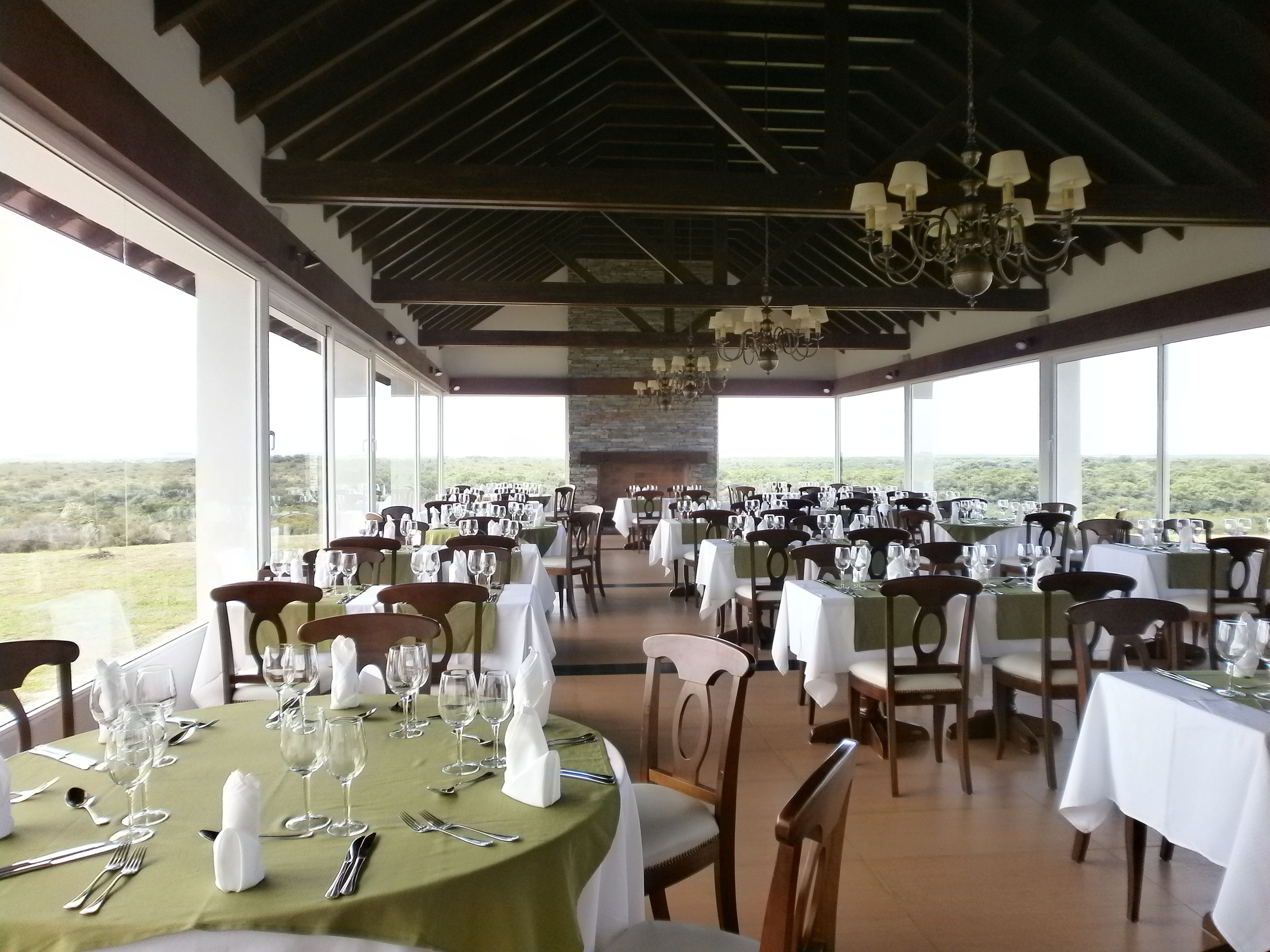 Un restaurante con varias mesas dispuestas para comer, amplias ventanas que ofrecen vistas del paisaje, y un candelabro colgando del techo.