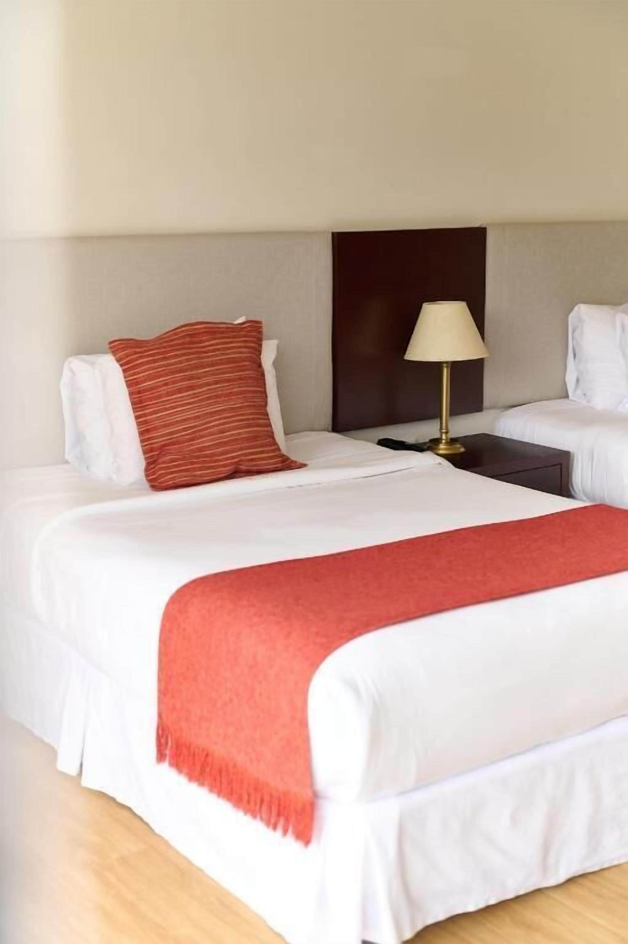 Una habitación de hotel con una cama grande, una mesita de noche, una lámpara y una manta roja.