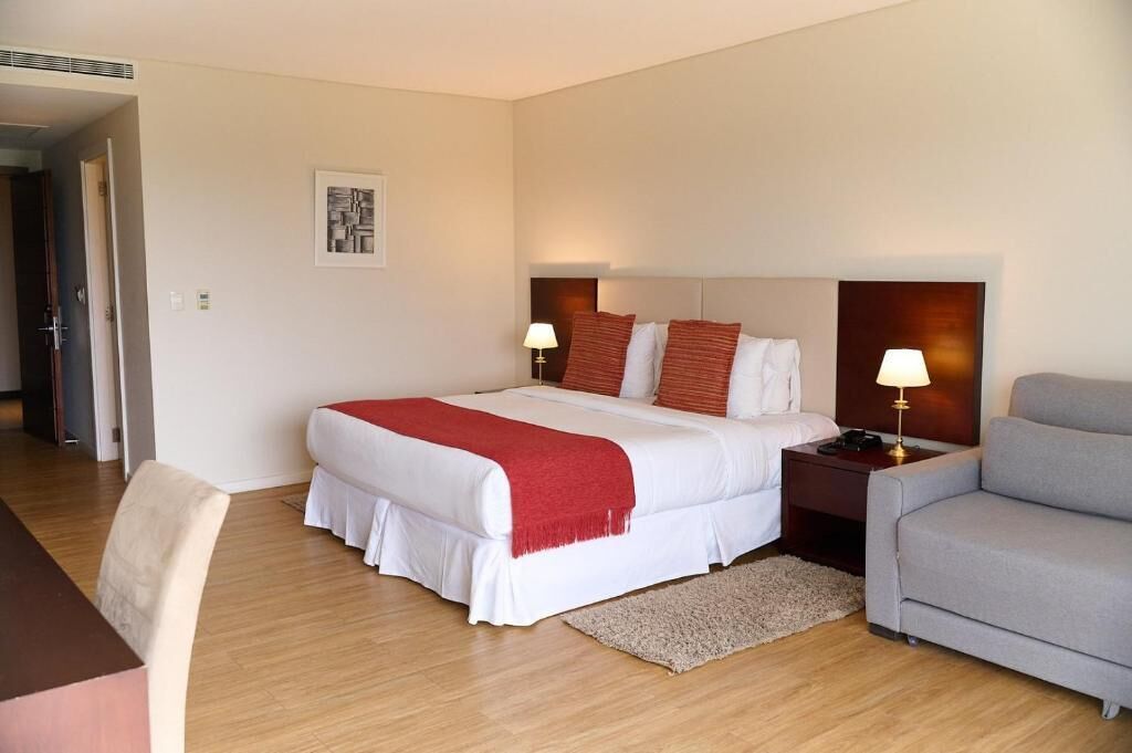 A hotel room with a large bed, a bedside table with a lamp, a grey armchair, and a wooden floor.