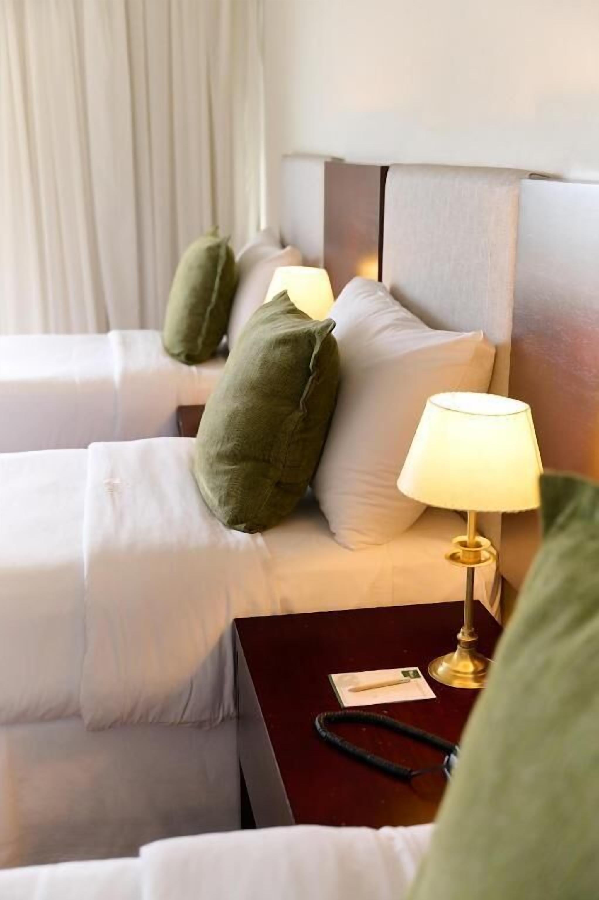 A hotel room with two beds, each with white linens and green pillows. There is a bedside table with a lamp and a black object on it.