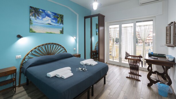 A bedroom with a bed, a desk, a chair, a large window, and a painting on the wall. - ViOletta (Rome)