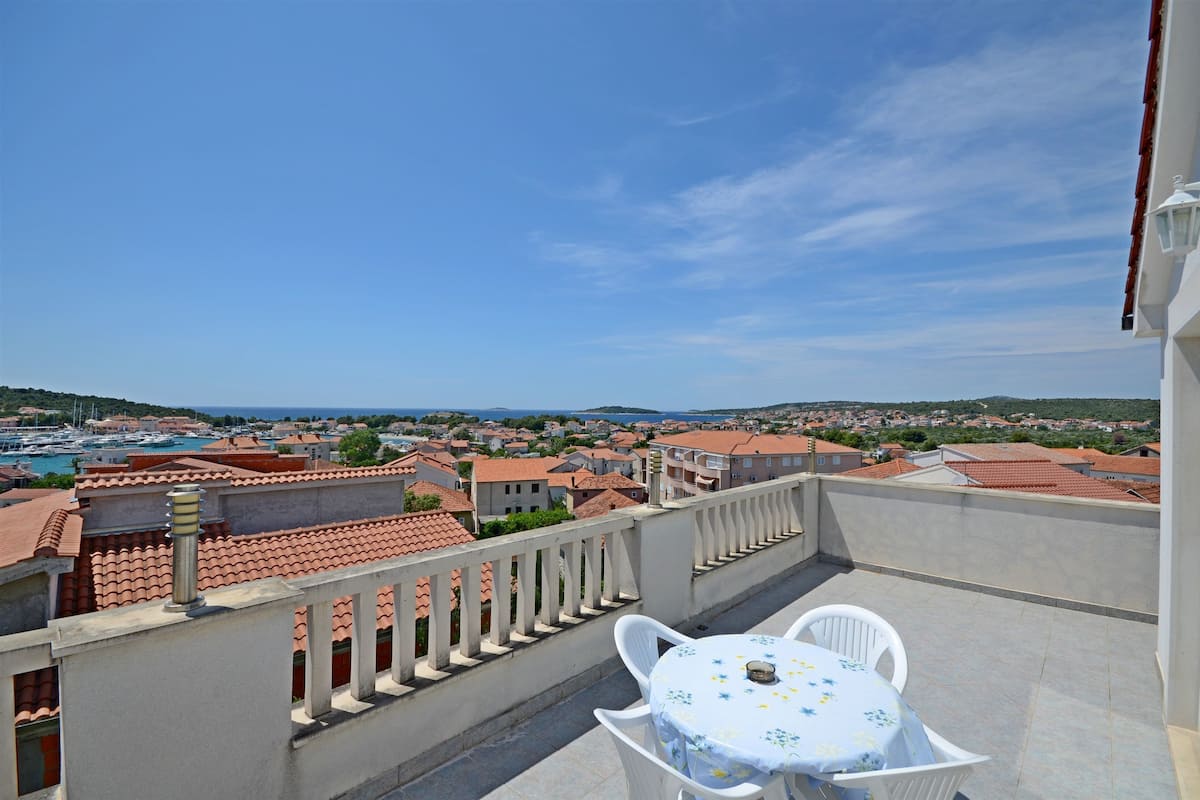 A balcony with a table and chairs, overlooking a coastal town with red-roofed buildings and a marina.