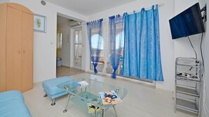 A room with a glass table, a blue sofa, a wooden wardrobe, and a TV mounted on the wall. - Apartments Branislav (Rogoznica)