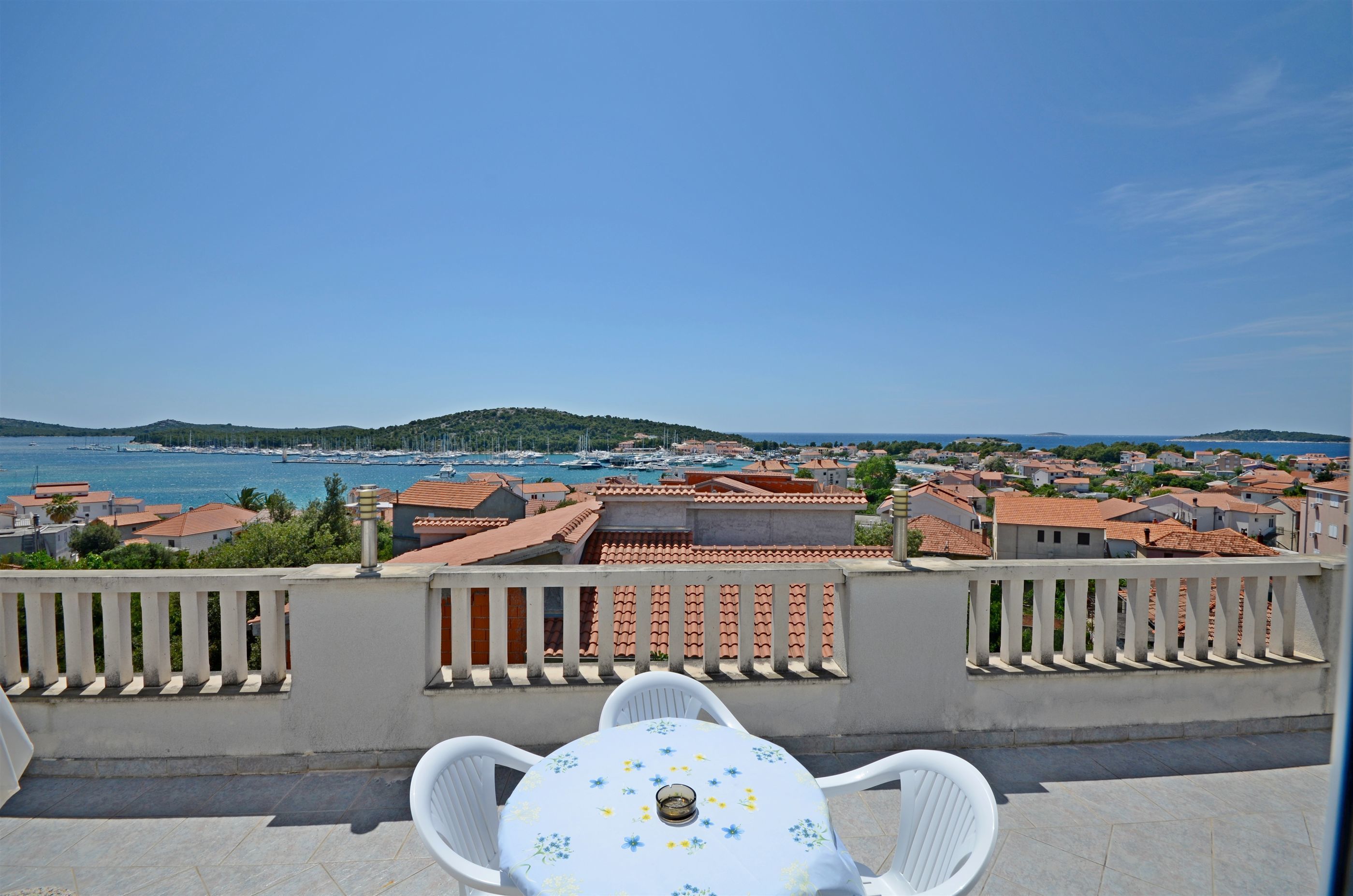 A terrace with a table and chairs overlooking a coastal town with red-roofed buildings and a marina.