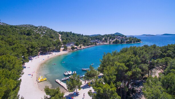 Une plage côtière aux eaux turquoise et limpides, bordée d’un rivage sablonneux, avec des collines verdoyantes en arrière-plan.
