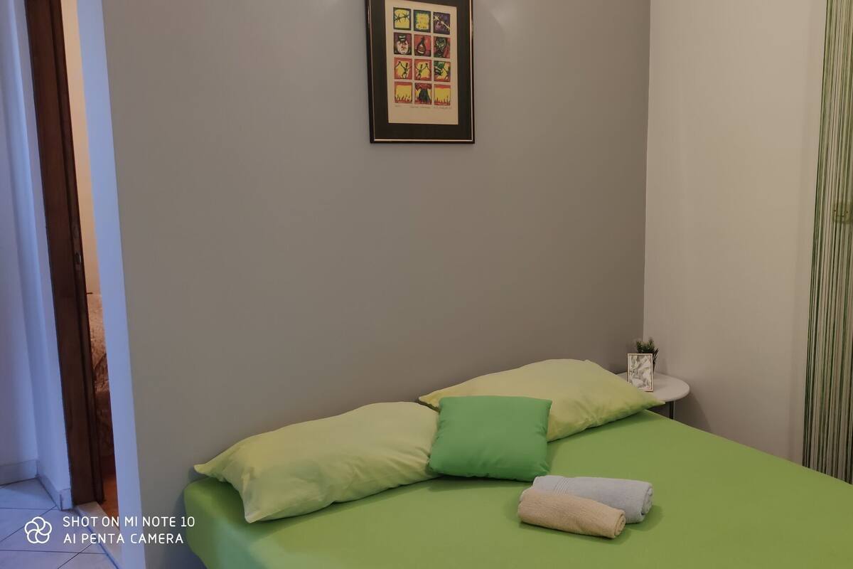 A room with a bed, pillows, a small table with a plant, and a framed picture on the wall.