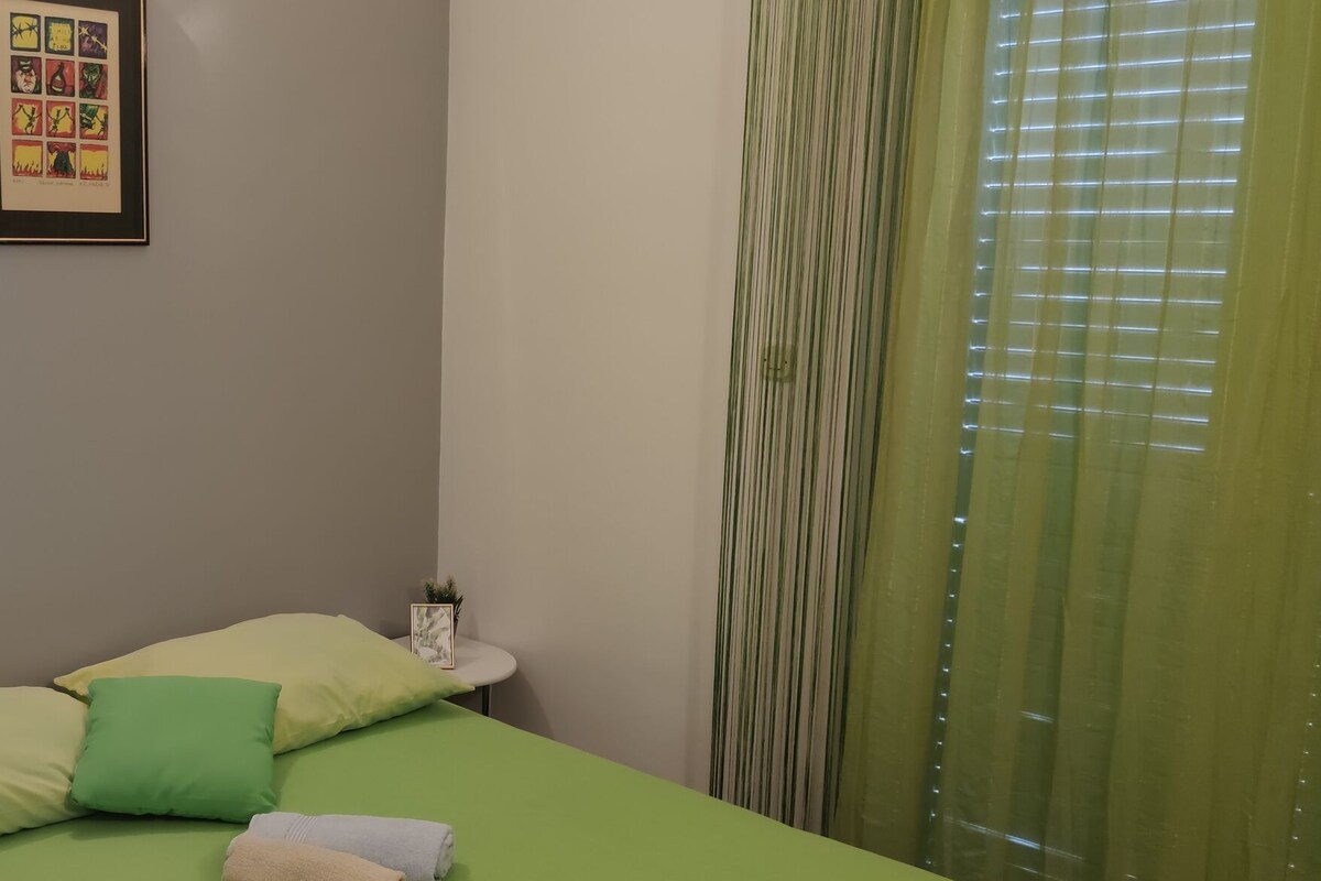 Ein Zimmer mit einem Bett, Kissen, einem kleinen Tisch mit einer Pflanze und einem gerahmten Bild an der Wand.