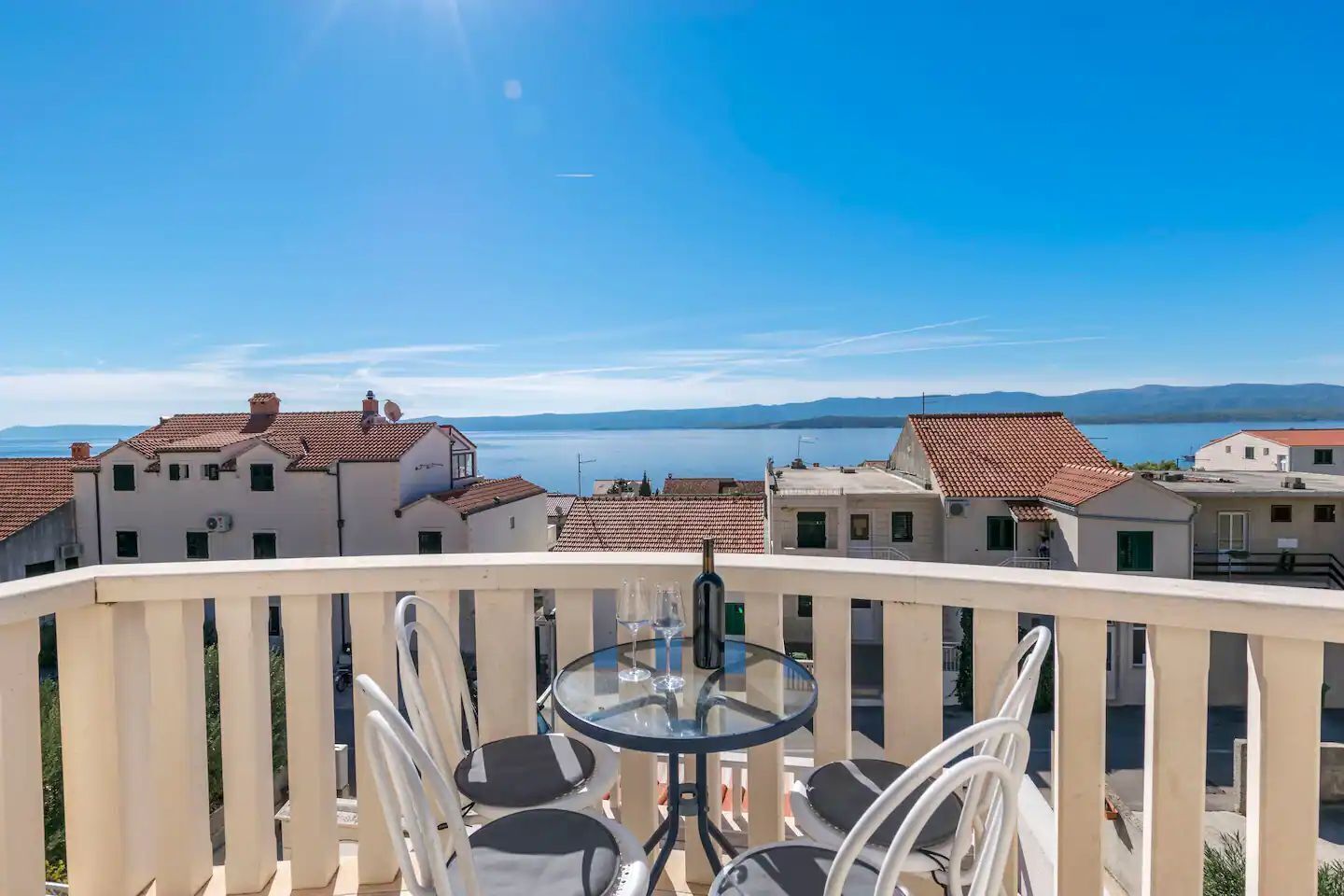 A balcony with a glass table and chairs, overlooking a coastal town with terracotta-roofed buildings and a clear blue sky.