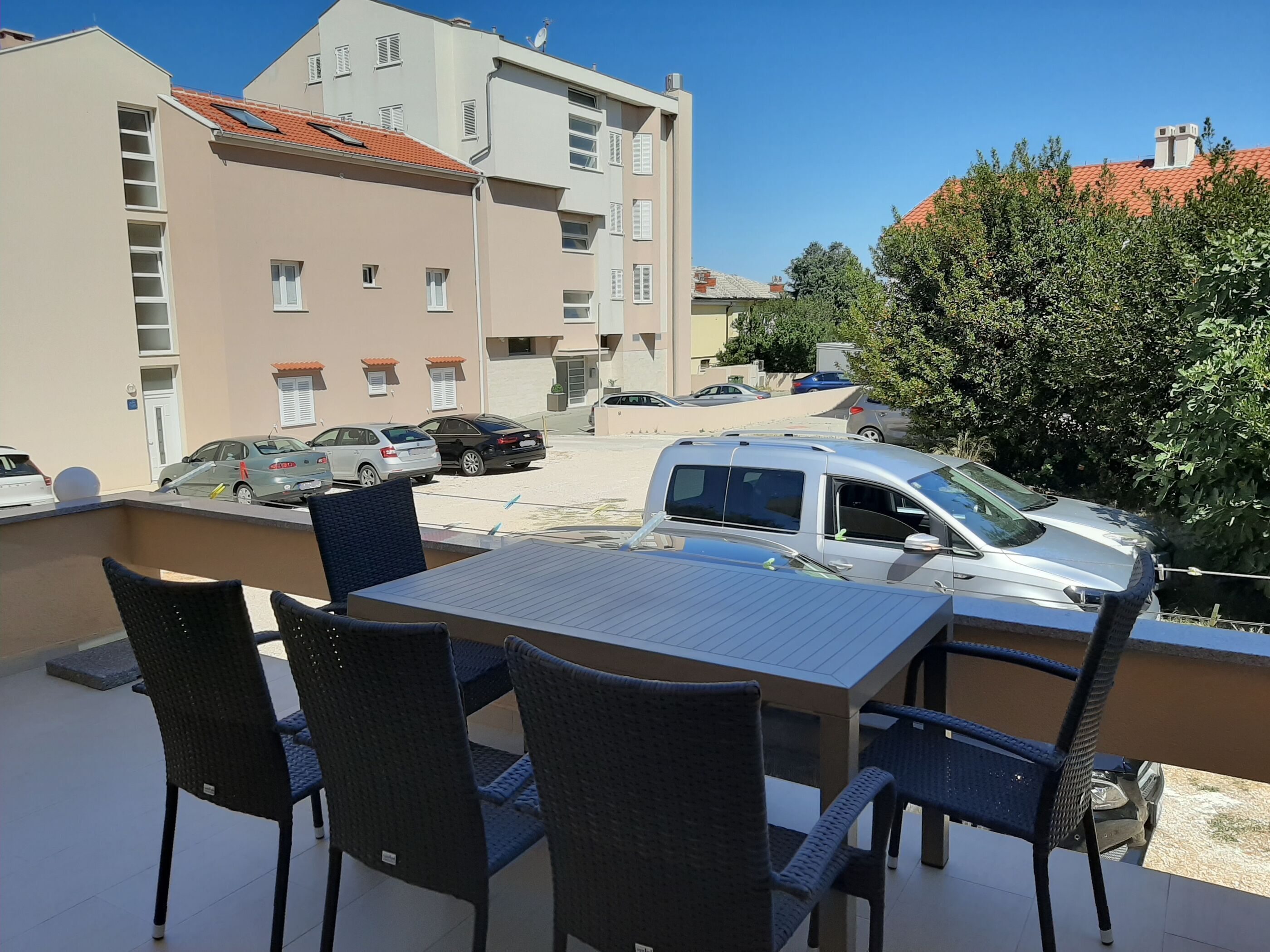 Balcón con mesa y sillas, con vistas a un estacionamiento con varios coches y un edificio con numerosas ventanas y balcones.