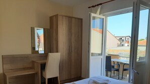 Un dormitorio con cama, escritorio de madera, silla, armario y balcón con mesa y sillas.