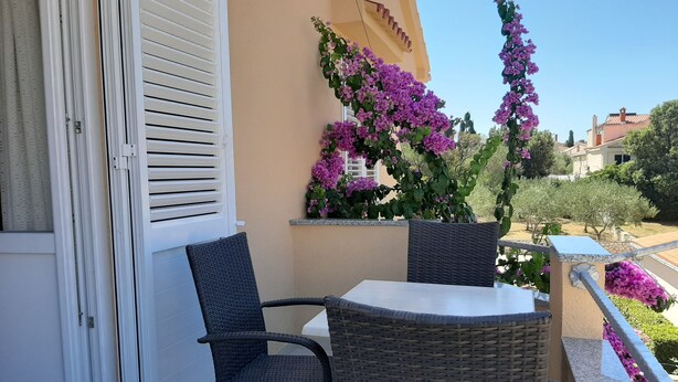 Un balcón con sillas de mimbre y una mesa, una ventana con persianas, y flores moradas.
