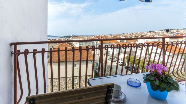 Un balcón con vista a un pueblo, una mesa con una taza y un platillo, y una planta en maceta.
