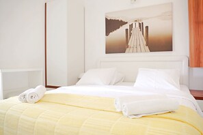Une chambre d’hôtel avec un lit, du linge de lit blanc, une couverture jaune et un tableau représentant une jetée accroché au mur.