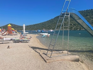 A beach with a playground, clear water, and boats. - House Suncani Dvori (Rogoznica)