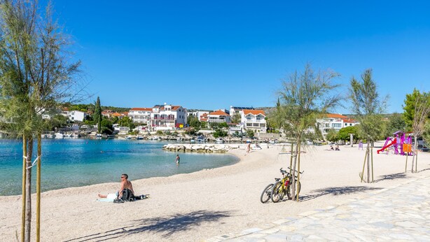 A beach with clear blue water, a sandy shore, and buildings in the background. - Apartments Luka (Rogoznica)