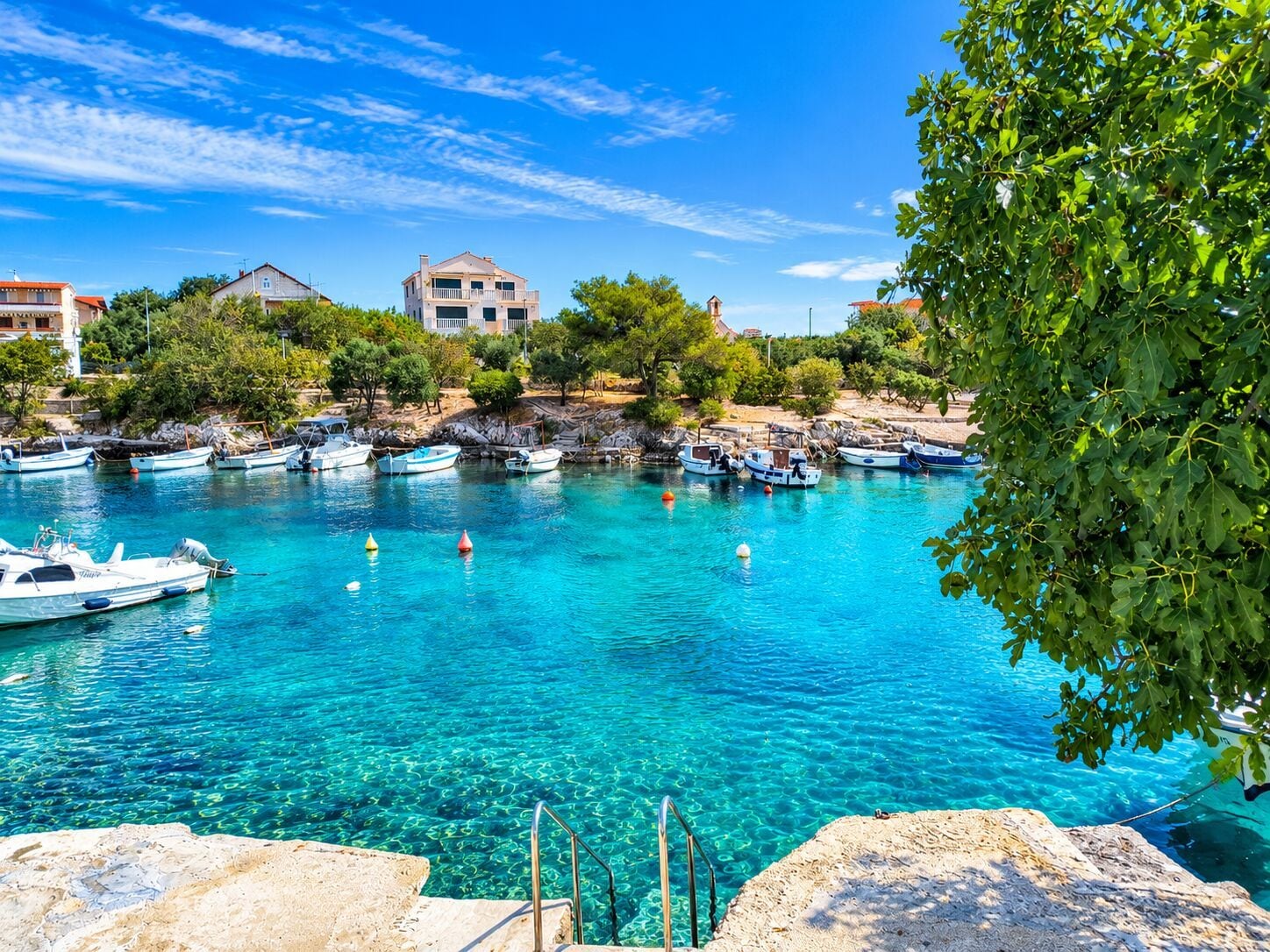 A coastal marina with boats docked, clear blue water, and buildings in the background.