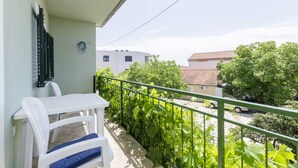 A balcony with a white table and chairs, a green railing, and a view of a residential area with trees and buildings. - Apartments Luka (Rogoznica)