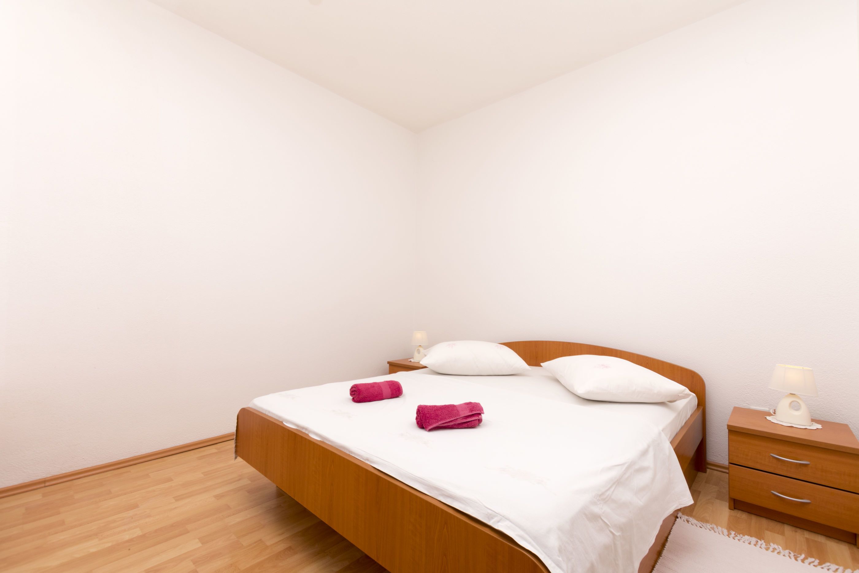 Une chambre avec un lit en bois, du linge de lit blanc, deux serviettes roses, une table de chevet avec une lampe et un plancher en bois.