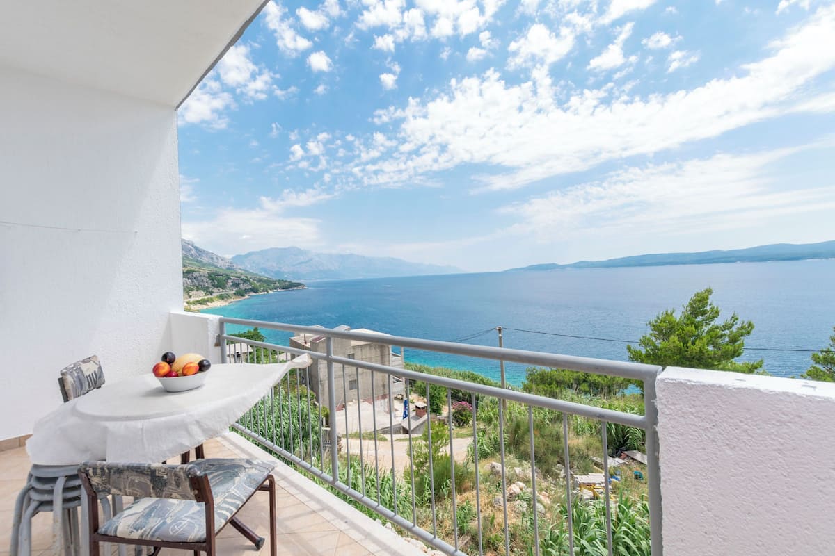 A balcony with a table and chairs, overlooking a coastal landscape with mountains and a body of water.