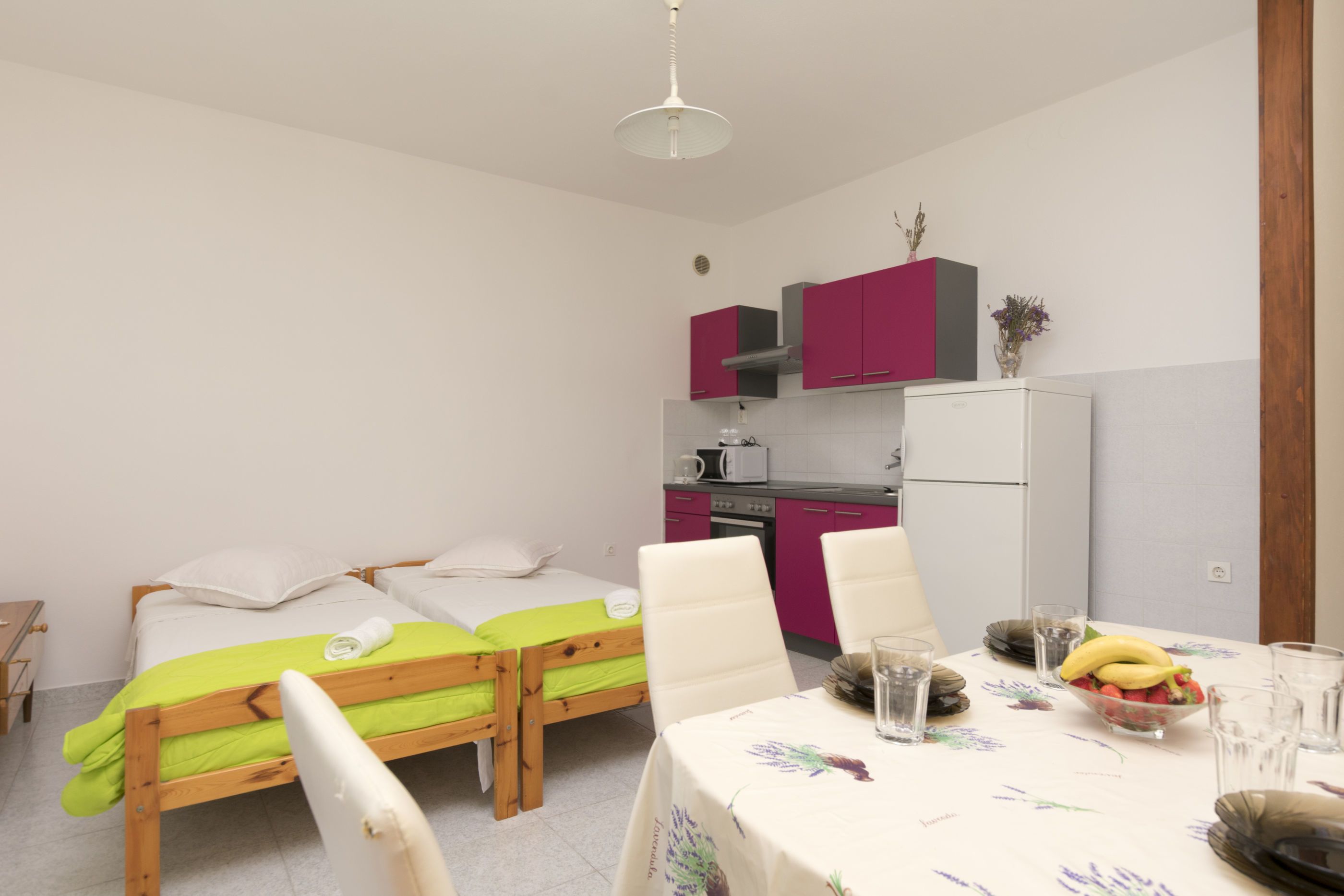 Una habitación pequeña y funcional con una cama, una mesa con mantel floreado y un rincón de cocina equipado con nevera y microondas.