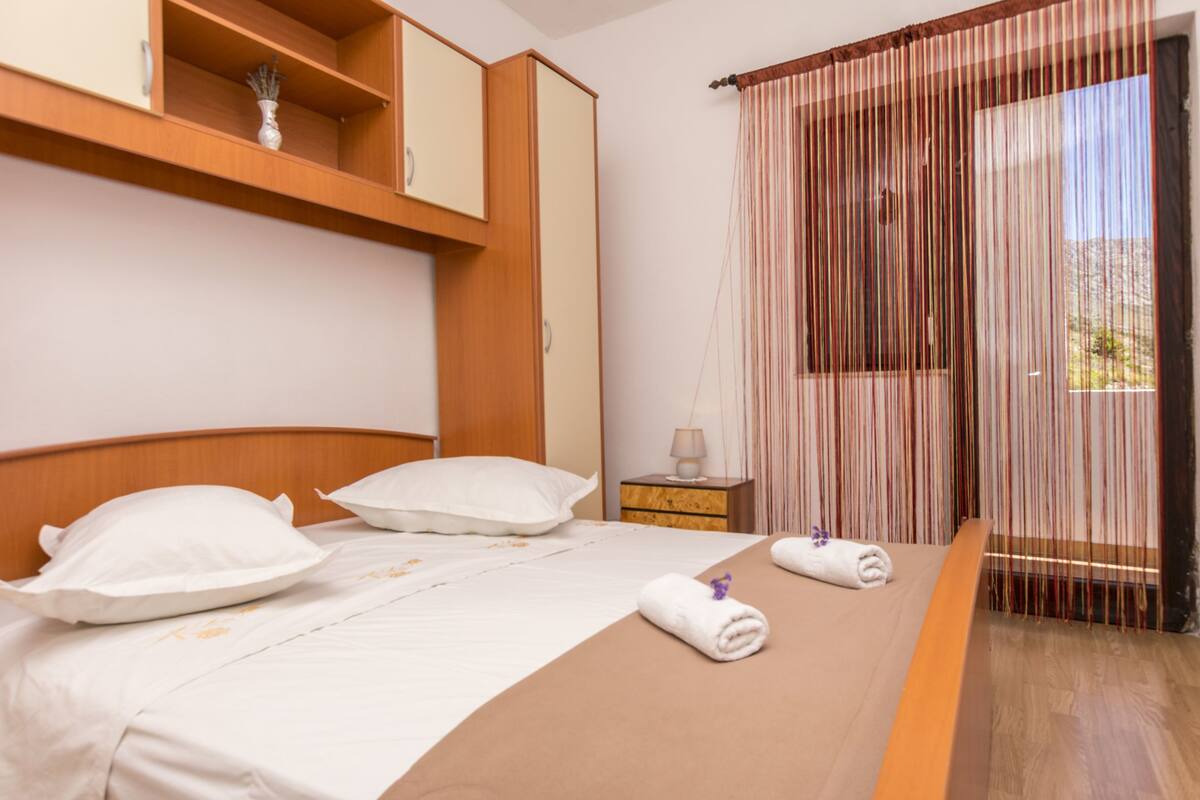 A neatly made bed with white linens and pillows, a wooden headboard, a bedside table with a lamp, and a window with vertical blinds.