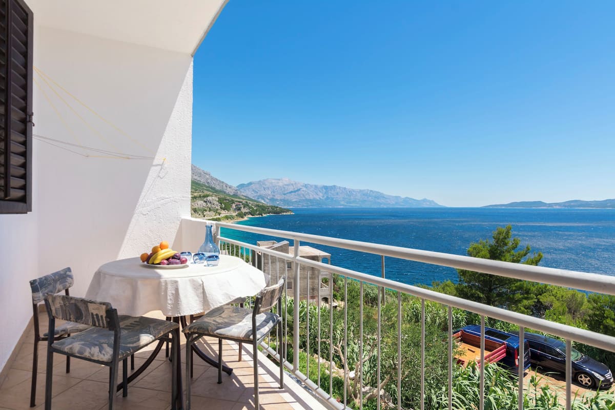 A balcony with a table and chairs, overlooking a coastal view with mountains in the distance.