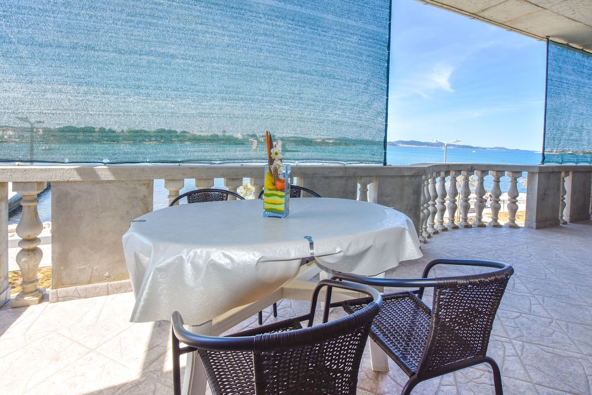 Ein überdachter Außenbereich mit Tisch und Stühlen, mit Blick auf ein Gewässer.