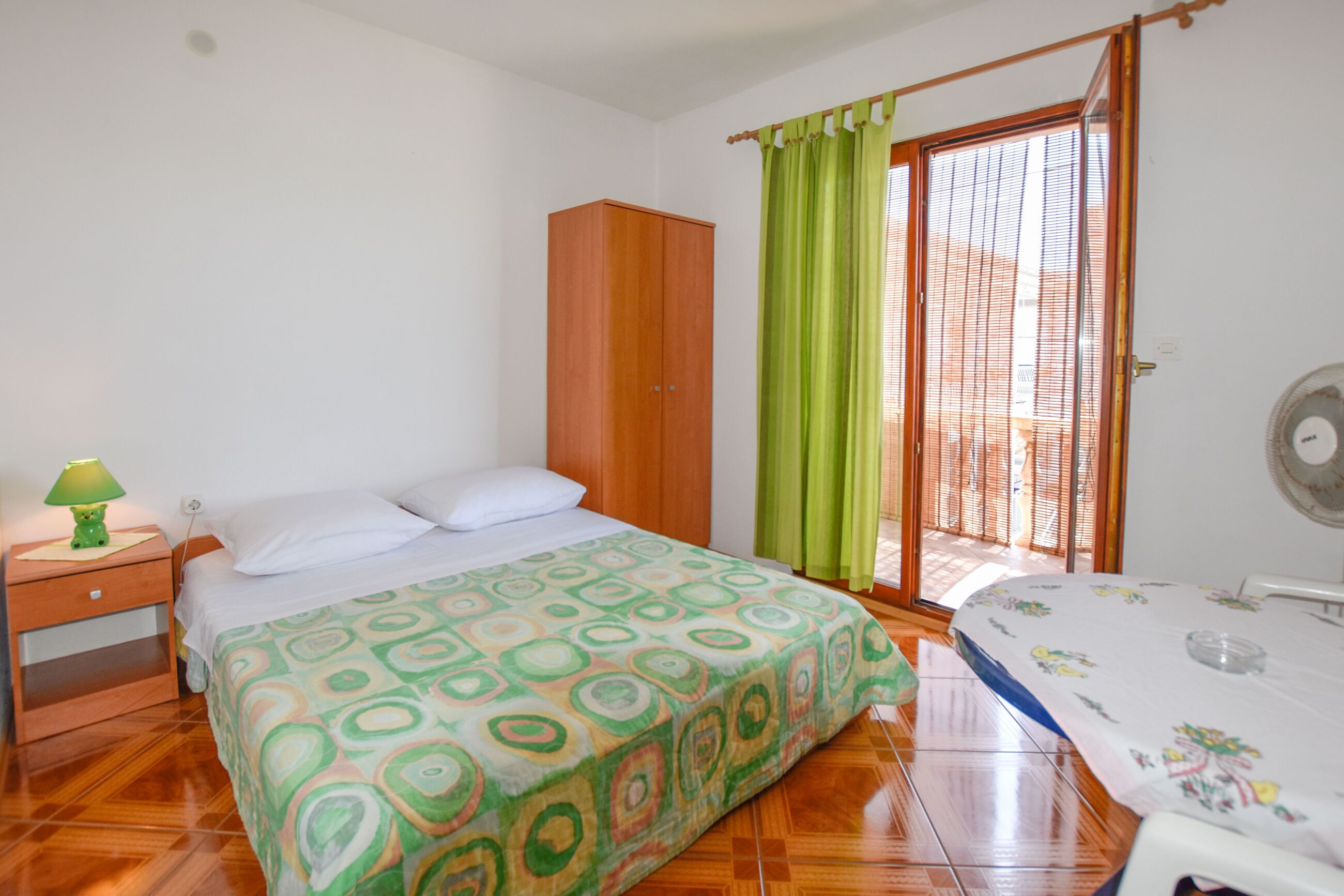 A bedroom with a bed, a nightstand, a wardrobe, a window with curtains, and a fan.