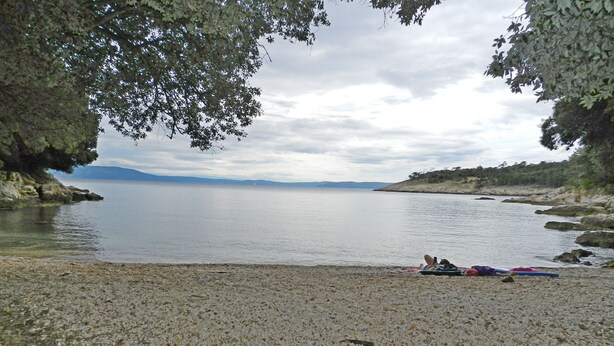 Une plage de galets bordant un lac paisible, avec un bateau et une personne allongée sur une couverture.
