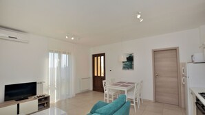 Un salon moderne comprenant un téléviseur à écran plat, un canapé, une table à manger avec des chaises, et un coin cuisine avec un réfrigérateur et une cuisinière.