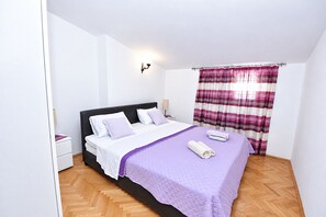 Egy egyszemélyes ágy fejvéggel, fehér ágyneművel és egy lila takaróval. Mellette egy éjjeliszekrény áll egy lámpával, az ablakon csíkos függöny van, a padló pedig fából készült.