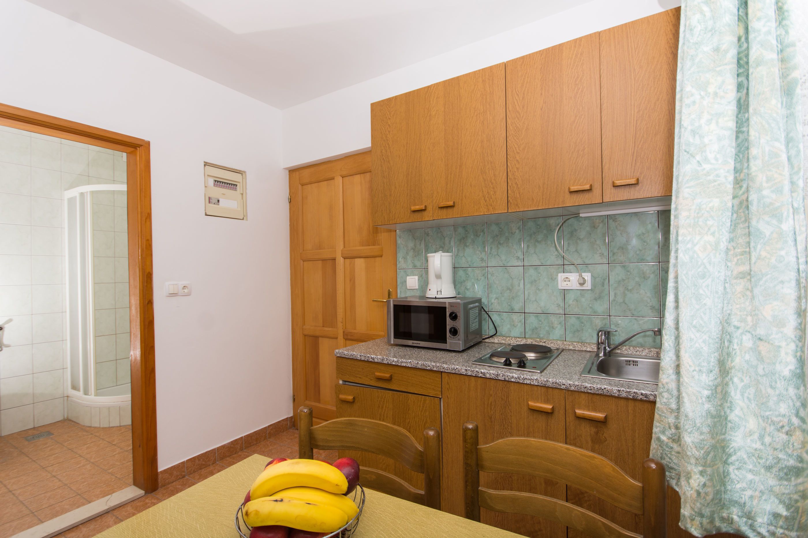 A compact kitchen with wooden cabinets, a microwave, and a sink. There is a shower visible in the background.