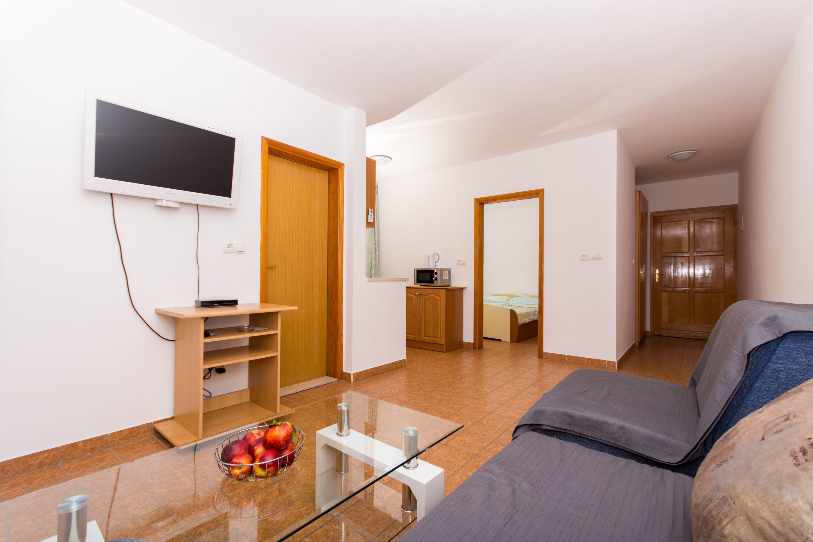Ein modernes Wohnzimmer mit Fernseher, einem Glastisch mit Früchten, einer Couch und einer Küche im Hintergrund.